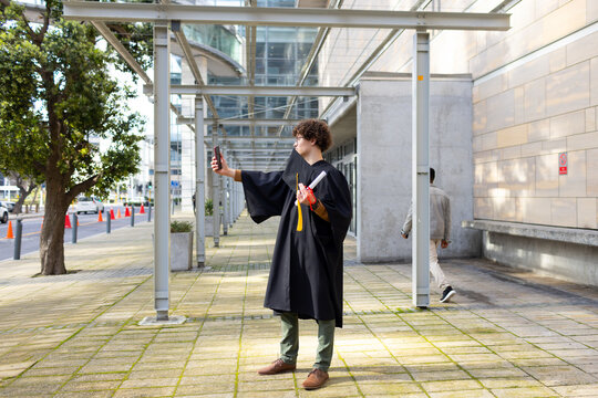 Male student wearing gown holding smartphone and diploma capturing selfie on walkway under canopy