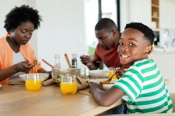 African American family sitting at wooden table in kitchen eating pasta with juice, copy space