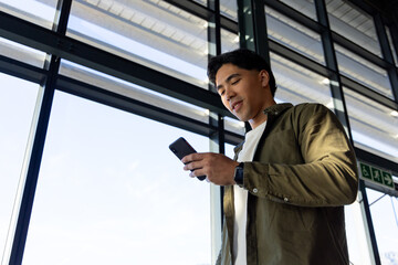 Man wearing olive shirt holding smartphone checking smartwatch near windows at airport, copy space