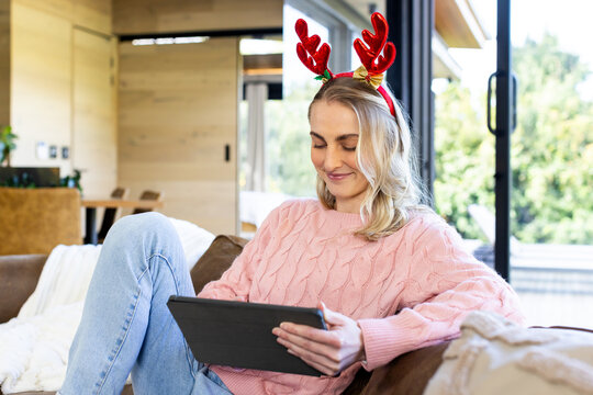 Woman sitting on brown couch holding tablet wearing red reindeer antler headband in living room