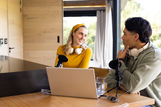 Podcast co-hosts recording episode at home studio desk with mics laptop headphones glass of water - Powered by Adobe