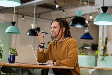 African American man speaking into smartphone at co-working table wearing brown sweater with laptop