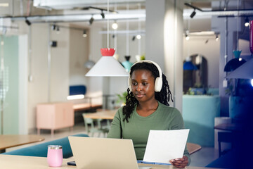 African woman sitting at shared space wearing green top, white headset, holding paper using laptop