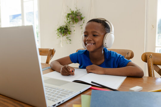 African American child sitting at dining table at home using laptop while wearing white headphones