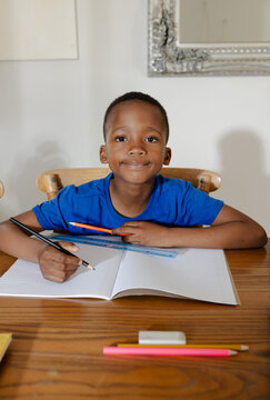 African American child sitting at home table wearing blue tee writing in notebook holding pencil