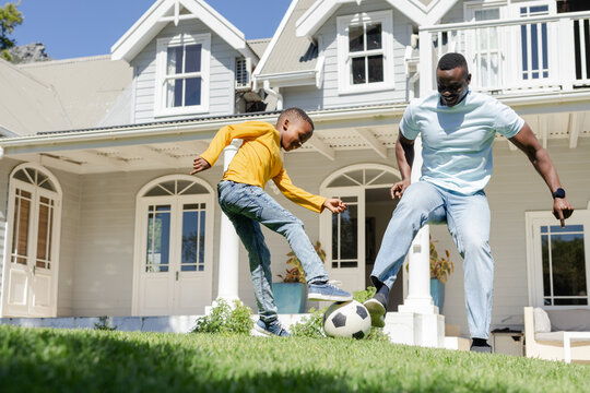 African American father and son kicking soccer ball on front yard wearing light-blue tee yellow top