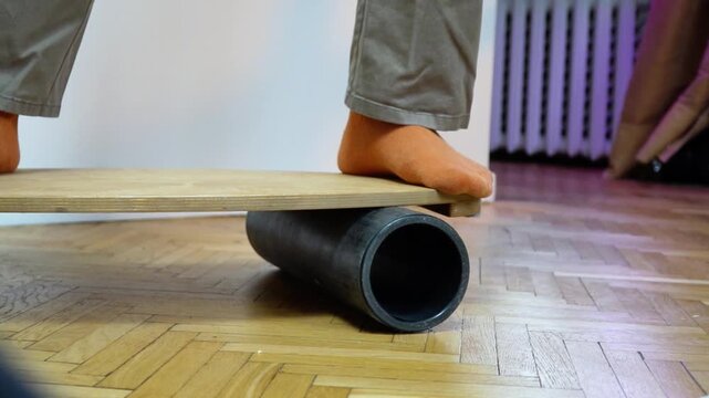 lowmotion barefoot person in casual pants balancing on wooden balance board with foam roller underneath training vestibular system and core stability indoors