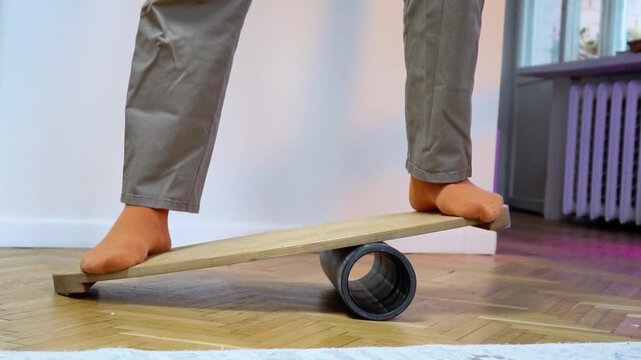 lowmotion barefoot person in casual pants balancing on wooden balance board with foam roller underneath training vestibular system and core stability indoors