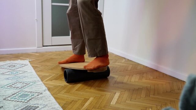 lowmotion barefoot person in casual pants balancing on wooden balance board with foam roller underneath training vestibular system and core stability indoors