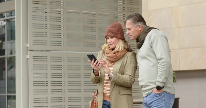 Senior couple lifting phone while leaning and tapping and scrolling screen sharing view on sidewalk