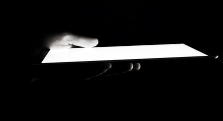 A hand holding a glowing tablet in the darkness. The bright screen contrasts with the black background, highlighting the technology and its usage
