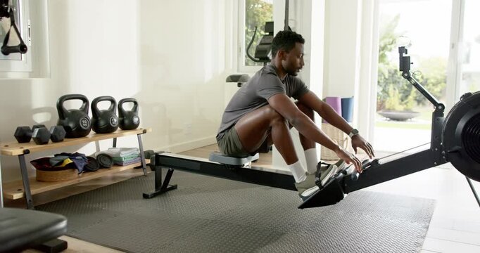 Mid adult African American man glancing at smartphone to track workout, rowing on machine at home