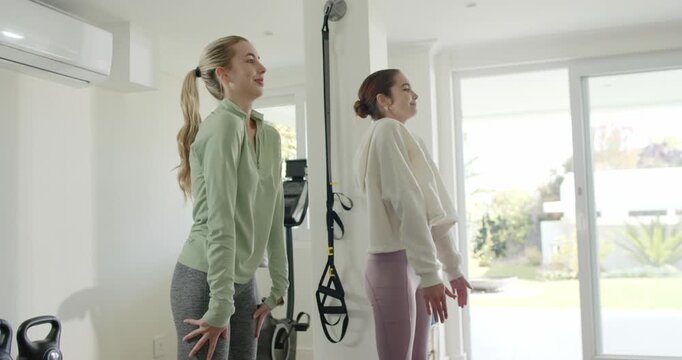 Two women shifting onto heels pushing hips forward for backbend stretch at gym with TRX straps