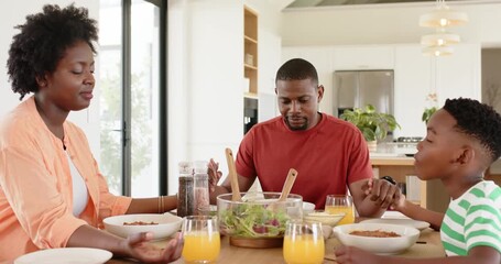 African American family holding hands praying before meal at home while son sliding juice to mother - Powered by Adobe