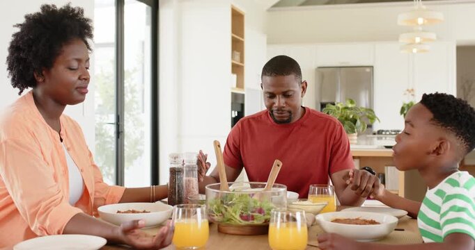 African American family holding hands praying before meal at home while son sliding juice to mother