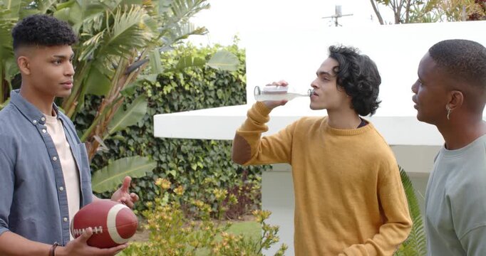 Diverse male friends standing yard shifting football explaining plan while sipping bottled beverage