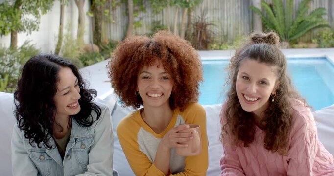 Diverse female friends glancing toward friend on sofa by blue pool, laughing, sipping copper mug