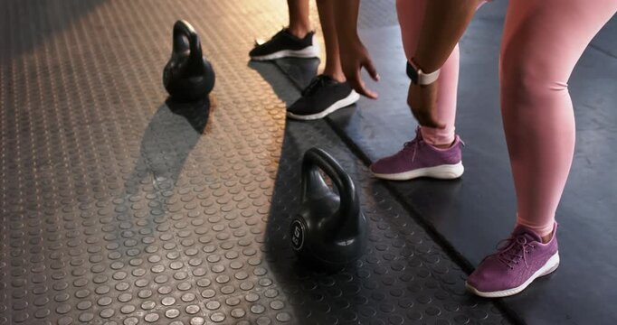 Diverse female friends gripping kettlebells, hinging and rising, bumping elbows and laughing at gym