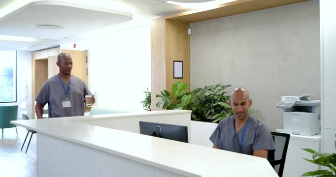 African American man in scrubs holding cup and wearing lanyard, entering, greeting coworker by desk