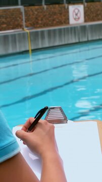 Vertical video: Timing coach in blue shirt at pool with pen on clipboard, timing laps at swim start