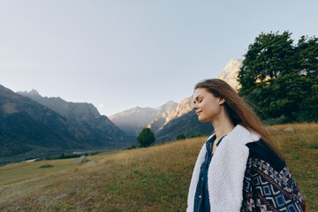 Naklejka premium Woman stands in meadow near mountains, portrait shot outdoors during sunrise, travel lifestyle scene showing peaceful landscape, young female in warm coat enjoying nature.