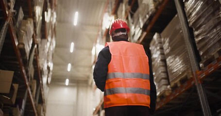 Warehouse worker in a high visibility vest and hard hat inspecting stock shelves, ensuring safety and organization in a large storage facility, low angle view highlighting warehouse scale.