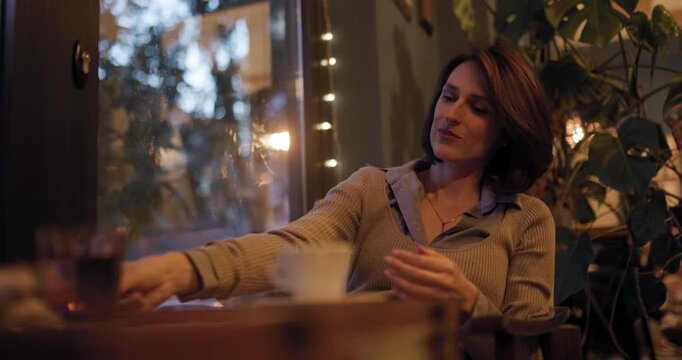 Woman in cozy cafe looking at smartphone with a slight smile, surrounded by warm lighting and greenery, enjoying a relaxing moment of leisure and connection.