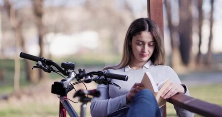 Captured in a serene park, this image shows a young woman reading a book with her bicycle nearby, enjoying a peaceful outdoor moment. - Powered by Adobe