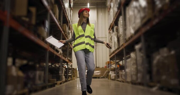 Warehouse worker confidently walking through aisle with a folder in hand, wearing a high-visibility vest and red hard hat, showcasing workplace efficiency and professionalism