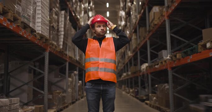 Warehouse worker in orange safety vest and red hard hat looking up in large industrial storage facility