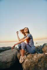 Lifestyle woman in modern boho-western denim street style sitting on rocks by the sea at sunset with film color warmth and relaxed pose