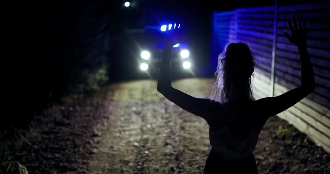 Girl with hands raised in surrender at night near police vehicle on a dirt road illuminated by flashing lights