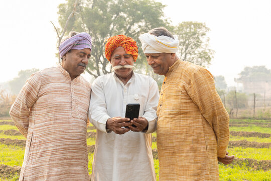 Three rural indian farmers using smart phone at agriculture field. Happy senior male friends check government scheme online on mobile phone. Wireless technology, Internet banking. Digital india
