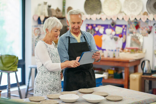 Creative, pottery and old couple with tablet, workshop and checking orders on website or production. Small business, elderly man and senior woman with tech for sales, craftsmanship or ceramic product - Powered by Adobe