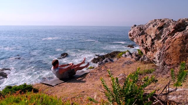 Man does seated abdominal plank hold core flutter kicks on sea cliff in morning
