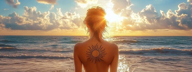 Person with a large sun tattoo on upper back standing topless facing the ocean during a golden sunset with clouds and gentle waves