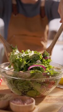 Woman preparing healthy green salad mixing with wooden spoons in glass bowl slow motion vertical video mindful cooking therapy clean eating home kitchen lifestyle