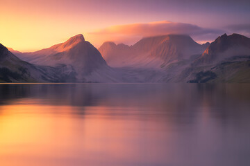 Stunning alpine lake reflects vibrant orange and purple sunset glow over majestic misty mountains at dawn