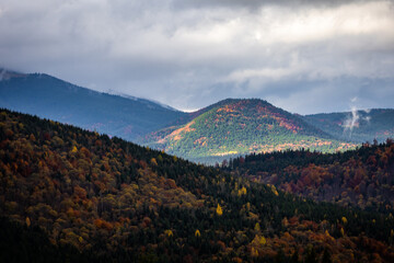 Sunlight breaks through heavy clouds to paint rolling mountain ridges with bands of autumn color and forested slopes fading to blue distances a dramatic and expansive landscape of wild highlands.
