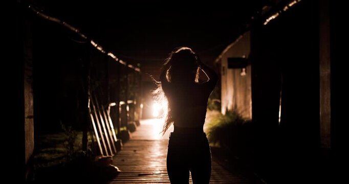 Girl in silhouette standing by car headlights at night with wind blowing through her hair in a quiet location