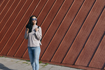 Stylish woman poses beside a diagonal wooden wall wearing sunglasses scarf and jeans showcasing...