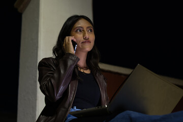 Young woman focused on her smartphone conversation and laptop activity at night, sitting outside, engaged in remote work, studying, or connecting with clients