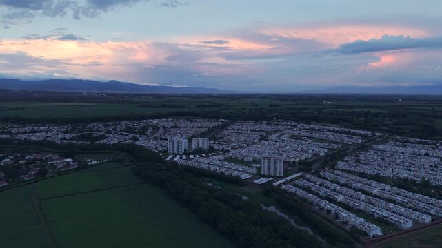 Drone video showcasing a scenic aerial view of a residential area in Valle del Cauca, Colombia, at sunset. Lush greenery surrounds neatly arranged homes under a colorful evening sky.
