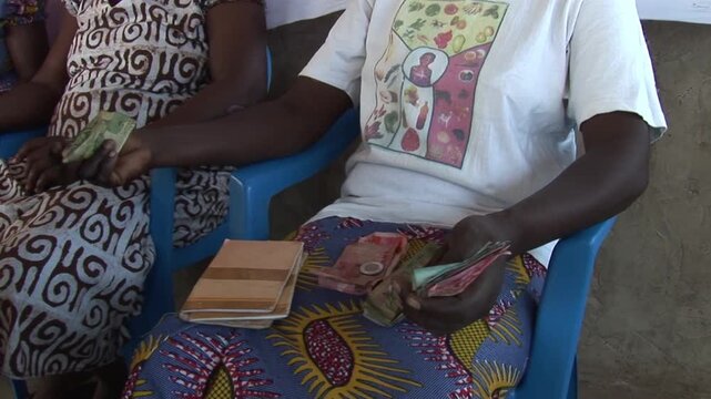 Women in a Ghanaian village savings and loan group sit together counting cash and holding passbooks during a microcredit meeting.