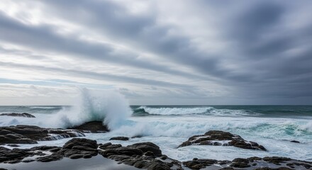 Obraz premium Turbulent ocean waves crash forcefully against dark coastal rocks beneath a sweeping cloudy sky