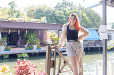 Asian beautiful woman orange long hair smile wear black top cozy cardigan light shorts, standing pose under warm sunlight calm waterside village charming riverside background travel relaxation holiday