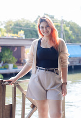 Asian beautiful woman orange long hair smile wear black top cozy cardigan light shorts, standing pose under warm sunlight calm waterside village charming riverside background travel relaxation holiday
