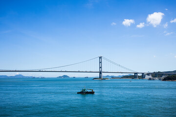 A suspension bridge over vibrant blue water with islands and ships in the distance under a clear sky.