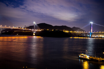 Nighttime view of two illuminated bridges over water, one cable stayed and one suspension, with reflections and city lights creating a vivid scene.