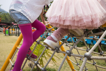 Two children climbing a rope net at a playground, dressed in bright clothes, surrounded by greenery and play structures.
